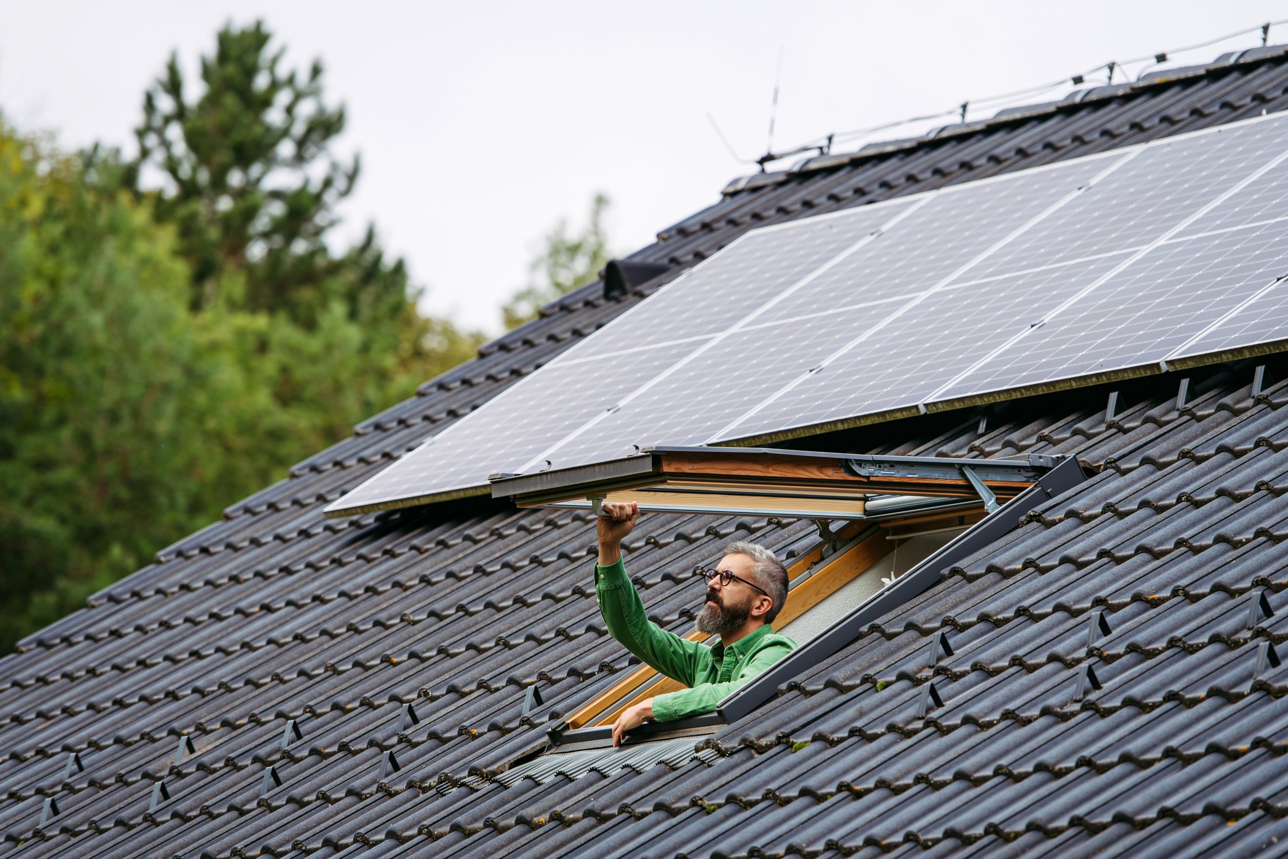 Mature man closing skylight, roof window, solar panels above him. Sustainable lifestyle and green household.
