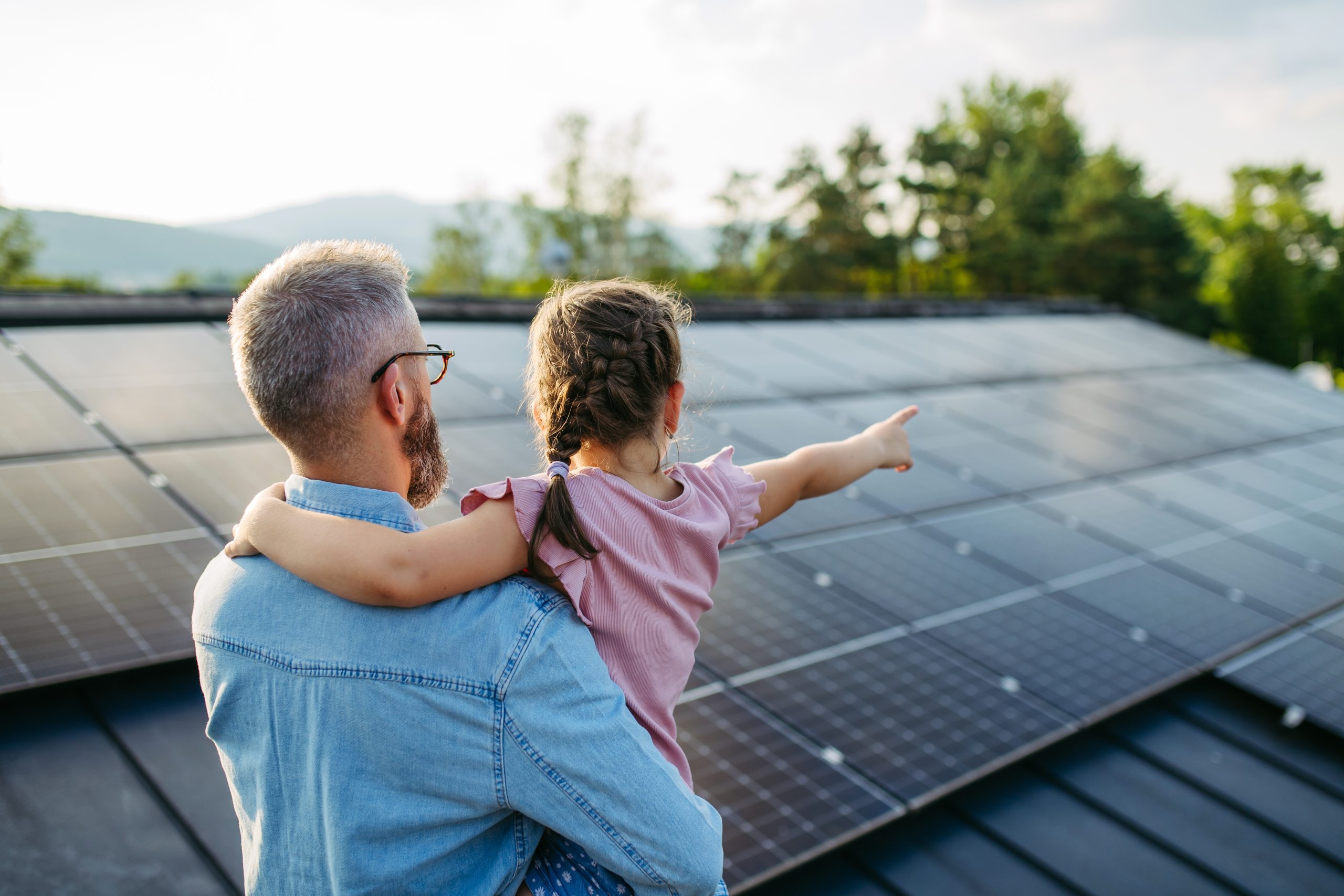 A father standing on the roof holding his daughter, who's pointing a finger at the installed solar home panels