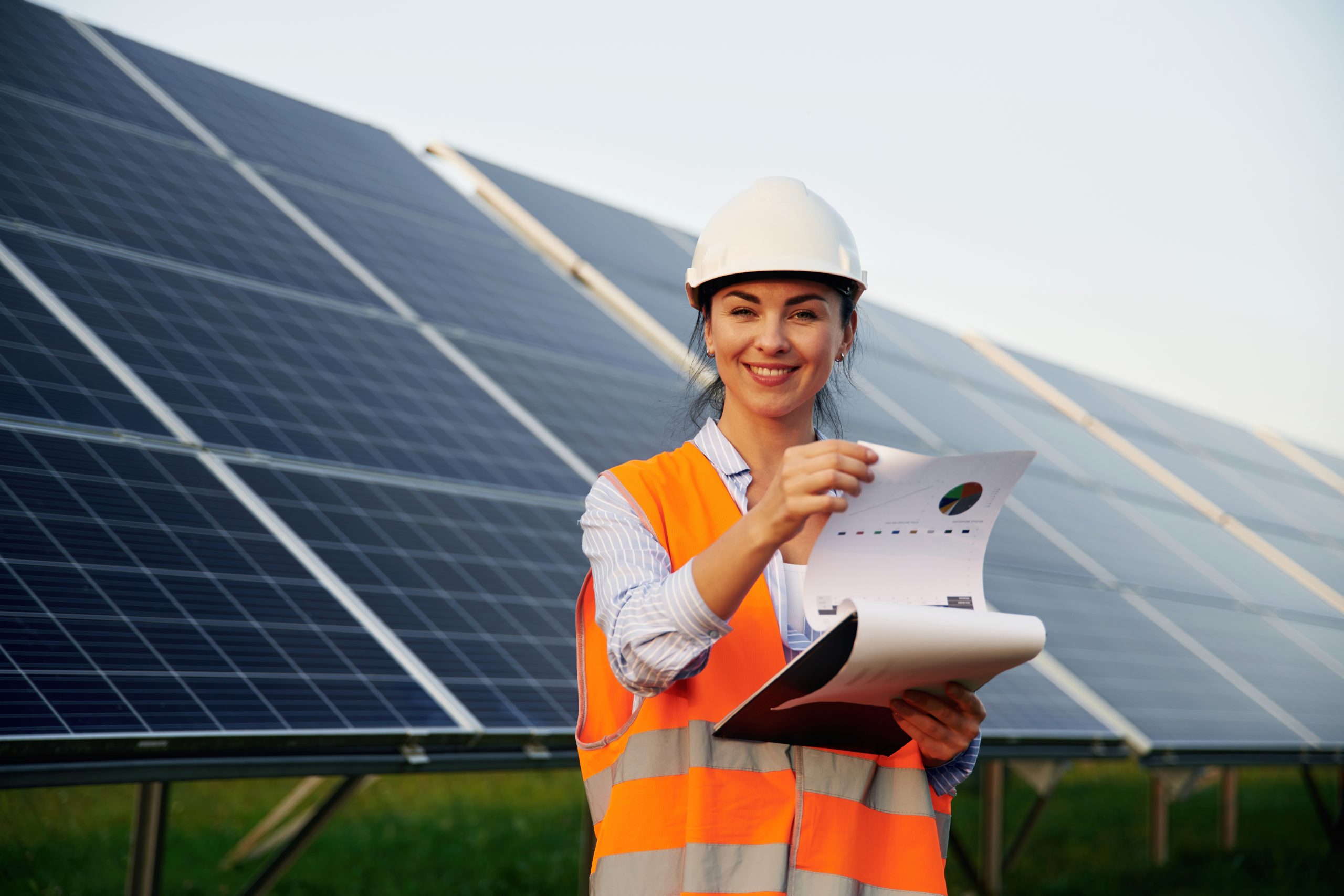 A technician in a hard hat and safety vest reviewing solar panel warranty documents while standing in front of ground-mounted solar panels.