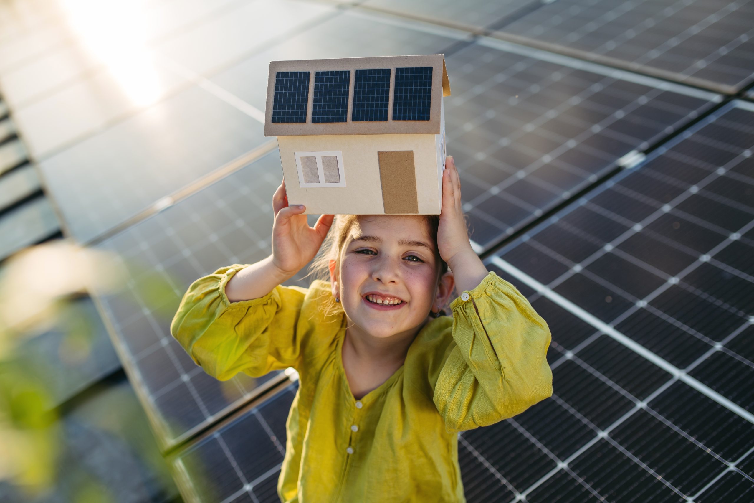 A little girl holding a model of a house with solar panels, sitting on a rooftop