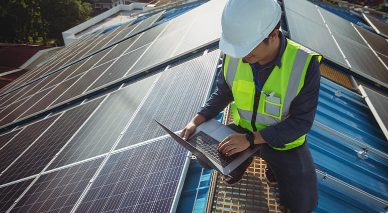 An engineer in a safety vest and hard hat inspecting solar panels while working on a laptop on a rooftop installation site.
