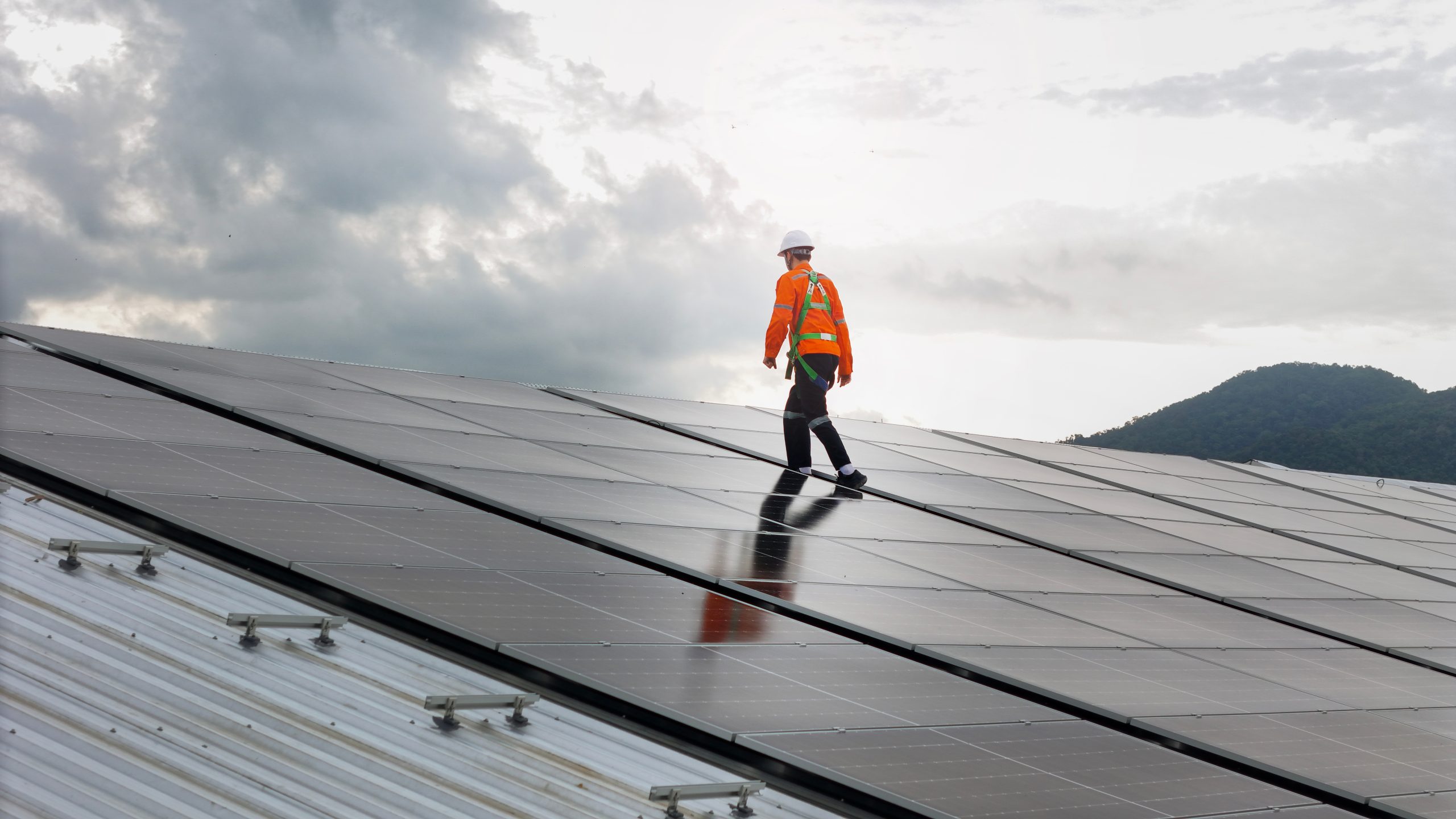 A worker checking solar panels on a rooftop installation under a cloudy sky.