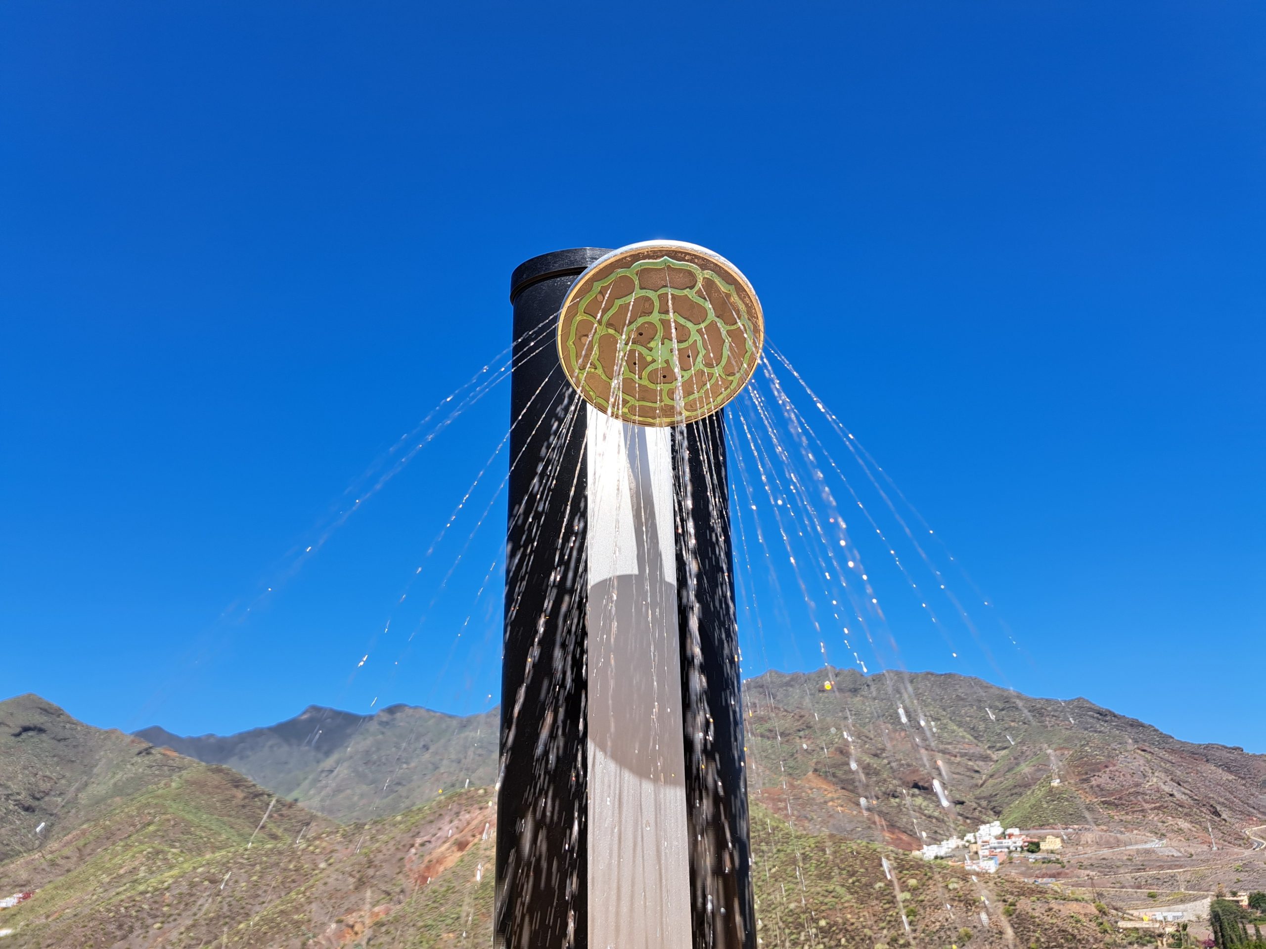 Solar shower spraying water outdoors against a clear blue sky with mountains in the background